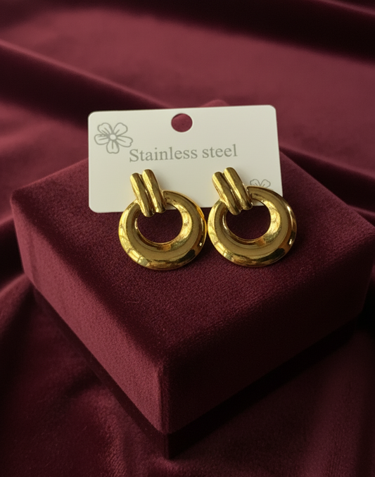 Gold earrings on a red cushion with a label indicating 'Stainless steel' on a red fabric background.